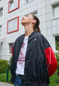 Černá a červená bomber bunda s lesklou texturou, která má na hrudi bílé logo. Pod ní se nachází bílá tričko s grafickým designem.