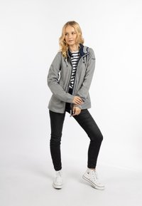 Giacca grigia con zip e cappuccio blu, dotata di cordino e tasche frontali, indossata sopra una maglietta a righe con jeans neri e sneaker bianche.