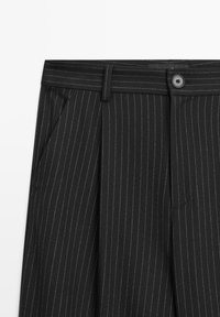 Schwarze Nadelstreifenhose mit plissiertem Design, ausgestattet mit einem Knopfverschluss und Seitentaschen. Der Stoff hat eine glatte Textur.