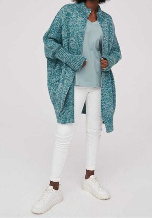 Personne portant un cardigan en tricot bleu sarcelle par-dessus une chemise bleu clair, un pantalon blanc ajusté et des baskets blanches, debout devant un fond uni.