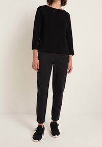 Svart långärmad topp, lös passform, i kombination med tighta svarta byxor och svarta sneakers med vita sålar, mot en ljus bakgrund.