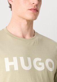 Alyvuogių žalios spalvos medvilninis marškinėliai su apvalia iškirpte, ant priekyje esantiems dideliems baltos spalvos užrašams "HUGO". Lygi tekstūra, laisvas fasonas.