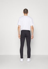 Jeans negros de corte ajustado con una textura suave, que presentan un diseño de cinco bolsillos y zapatillas blancas, mostrados desde la parte trasera sobre un fondo liso.