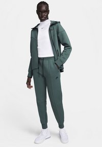Groene hoodie met zijknopen en zwart Nike-logo, gecombineerd met bijpassende joggers, heeft een elastische tailleband en manchetten aan de enkels, gedragen met witte sneakers.
