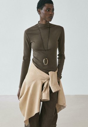 Kvinde iført brun langærmet top og bukser med beige jakke bundet om livet, accessoriseret med en lang oval vedhængshalskæde.