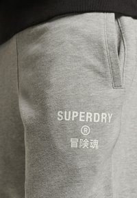 Sive trenerke napravljene od mekanog materijala s bočnim džepovima. Sadrži bijeli natpis "SUPERDRY" na butini u jednostavnom fontu.