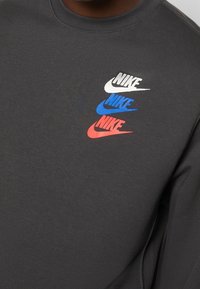 Mörkgrå tröja med tre lager av Nike swoosh-loggor i vitt, blått och rött. Tillverkad av mjukt, slätt tyg med design för rund hals.