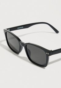 Μαύρα πλαστικά γυαλιά ηλίου με σκοτεινούς φακούς, τετραγωνικό σχήμα και διακριτικές ασημένιες λεπτομέρειες στο σκελετό. Σχεδιασμένα με κομψό, μοντέρνο ύφος.