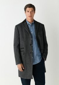 Sötét szürke gyapjú keverék kabát, túlórás gallérral, négy gombbal és két elülső zsebből áll, mely egy világoskék gombos ing felett viselendő.