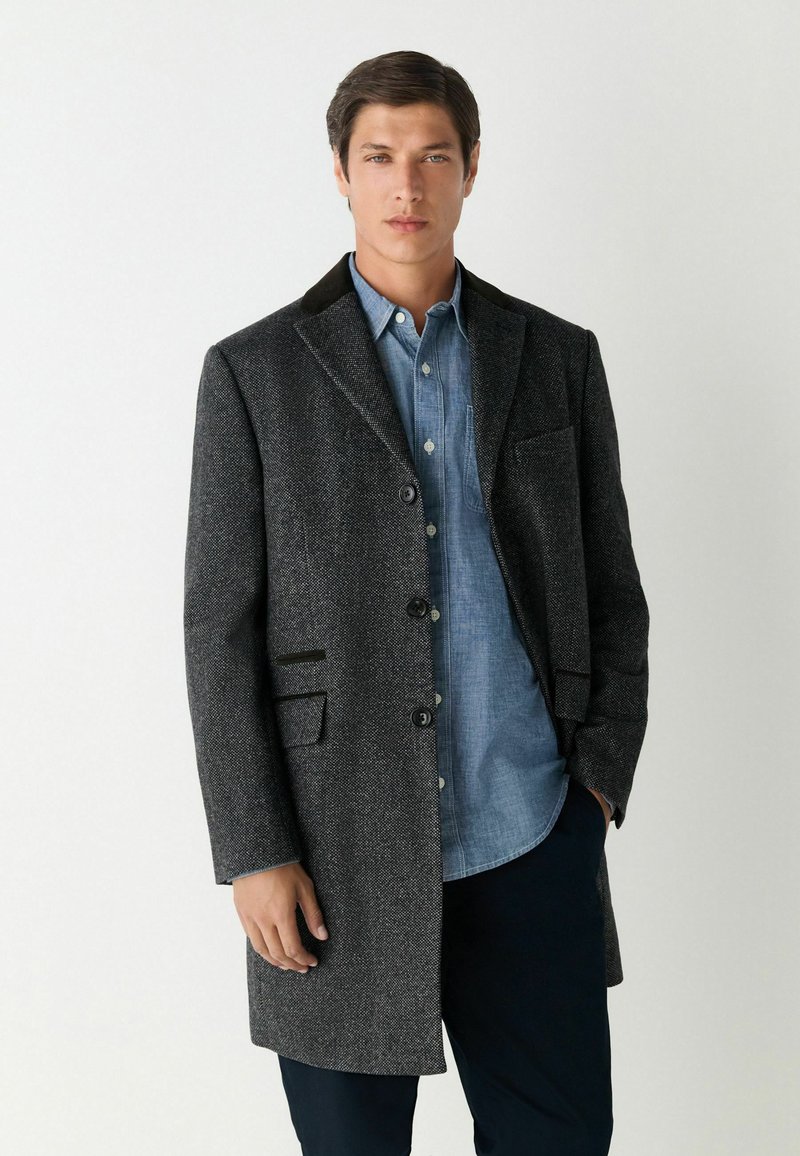 Sötét szürke gyapjú keverék kabát, túlórás gallérral, négy gombbal és két elülső zsebből áll, mely egy világoskék gombos ing felett viselendő.