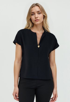 Top nero a maniche corte con scollo a V, caratterizzato da un accento dorato al centro. Tessuto liscio e vestibilità comoda, abbinato a pantaloni neri.
