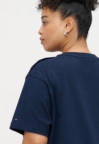 Navyblaues Baumwoll-T-Shirt mit kurzen Ärmeln, das über Schulterbeschläge und einen kleinen Logoakzent am Ärmel verfügt. Glatte Textur.