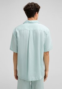 Himmelblaues Kurzarmhemd mit spitzem Kragen, lockerer Passform und glatter Textur. Mit einer Rückensattelnaht und dezenten Falten.