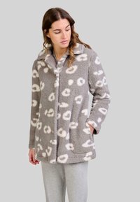 Mujer con una chaqueta de forro polar gris con manchas de leopardo blancas, de pie con una mano en el bolsillo, mirando hacia abajo a un lado.