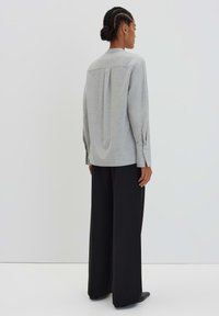 Chemise grise à manches longues avec un pli au dos, ourlet arrondi et poignets boutonnés, associée à un pantalon noir ample. Texture lisse, design minimal.