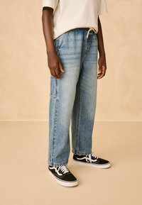 Pantaloni cargo din denim de culoare albastru deschis, cu o croială lejeră, având buzunare laterale și un talie cu șnur. Asortați cu adidași negri.