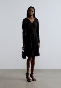 Must pikkade varrukatega kleit V-kaelusega ja keha kujundav paigutus, mis lehvib vööst allpool. Komplektis mustad rihmikud kõrged kontsad ja väike käekott.