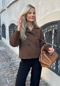 Brown double-breasted jacket with large buttons, worn over a dark outfit, paired with a brown handbag. Smooth texture; casual fit.