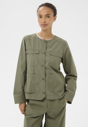Woman wearing olive green buttoned utility jacket with four large pockets and matching pants, standing against a plain white background.
