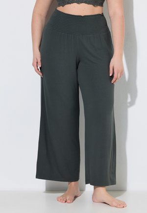 Donna che indossa pantaloni neri larghi e morbidi con vita elasticizzata, in piedi a piedi nudi contro un muro bianco.