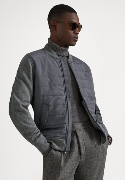 Szara pikowana kurtka z ciemnoszarymi rękawami, zapinana na zamek z przodu oraz z bocznymi kieszeniami, zestawiona z szarym golfem i dopasowanymi szarymi spodniami.