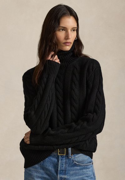 Jeune femme aux longs cheveux foncés portant un pull à col roulé en maille torsadée noire et un jean bleu, regardant sur le côté avec une expression neutre.