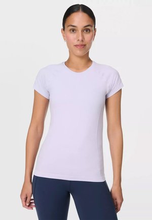 Young woman with pulled-back hair wearing a fitted light purple short-sleeve shirt and dark blue pants, standing against a plain background.