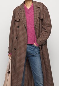 Brun och svart rutigt långkappa med en avslappnad passform, lager på lager över en livlig rosa tröja, tillsammans med blå jeans och en ljus handväska.