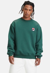 Felpa verde con collo a girocollo a costine, spalle abbassate e una piccola toppa loggata ricamata sul petto. Tessuto texturizzato, vestibilità comoda.