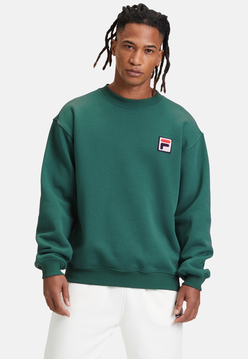Felpa verde con collo a girocollo a costine, spalle abbassate e una piccola toppa loggata ricamata sul petto. Tessuto texturizzato, vestibilità comoda.