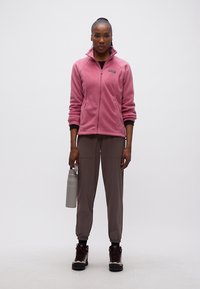 Γυναίκα στέκεται προς τα εμπρός φορώντας ροζ φλις μπουφάν με φερμουάρ, παντελόνια χρώματος ταουπέ, μαύρες κάλτσες, παπούτσια πεζοπορίας, κρατώντας ένα γκρι παγούρι νερού.