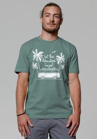 Groene t-shirt met witte grafiek van een bus met palmbomen en de tekst "Laat het avontuur beginnen!" Zacht materiaal, ronde hals ontwerp.