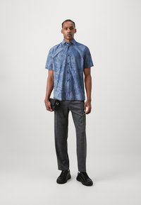 Camicia blu a maniche corte con motivo a foglie, pantaloni grigi con motivo, sneakers nere, tiene in mano una borsa nera con dettaglio ad anello.