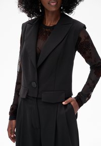 Czarna krótka kamizelka z wyłogowym kołnierzem, jednopunktowym zapięciem na guzik i przednimi kieszeniami. Nosi się na przezroczystą, kwiecistą bluzkę z długim rękawem.