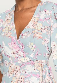 Květinové šaty s zavinovacím designem vyrobené z světle modré látky, zdobené růžovými a žlutými květy, s výstřihem do V a krátkými rukávy, měkkou texturou a výrazným pasem.