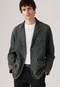 Joven vestido con un blazer de mezclilla oscura descolorido sobre una camiseta blanca y pantalones negros, de pie con las manos en los bolsillos contra un fondo simple.