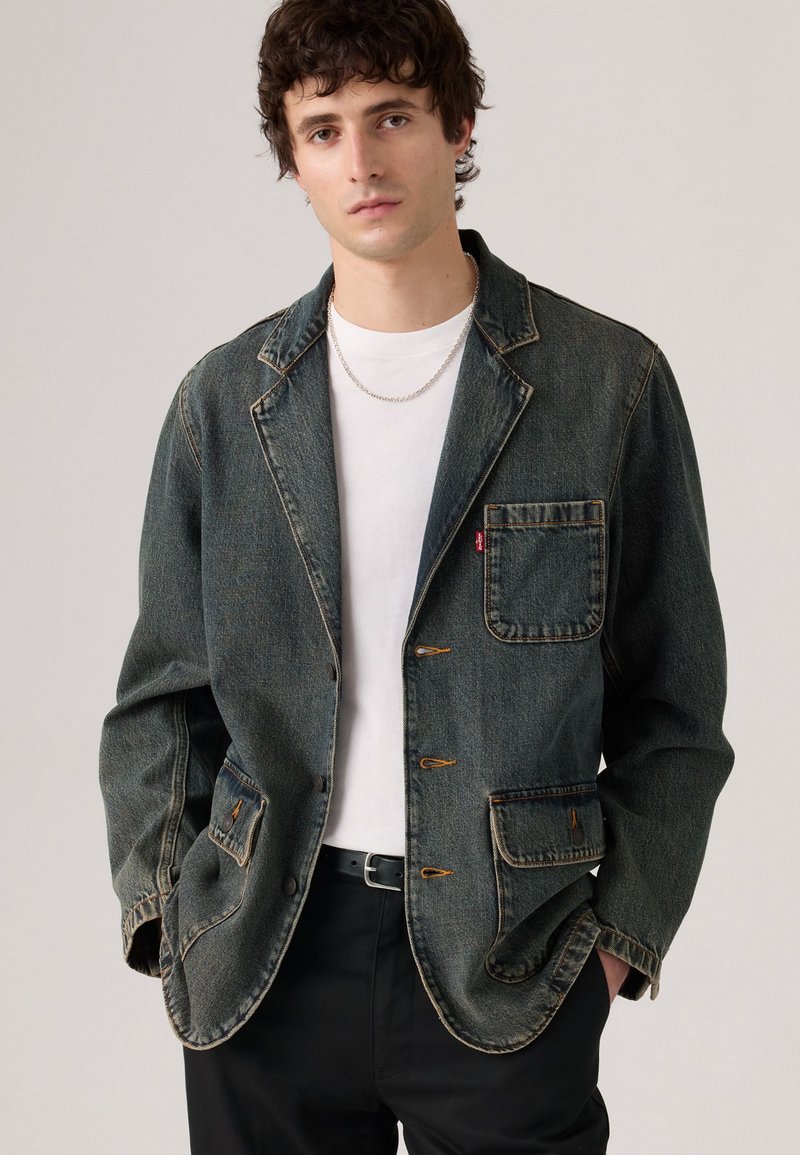 Joven vestido con un blazer de mezclilla oscura descolorido sobre una camiseta blanca y pantalones negros, de pie con las manos en los bolsillos contra un fondo simple.