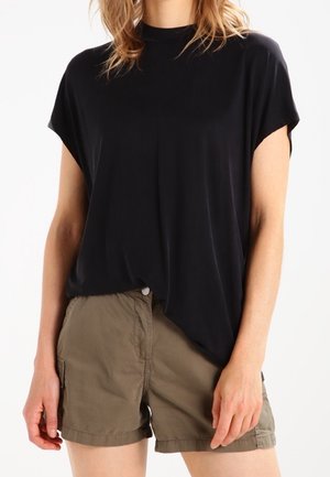 Vrouw draagt een losse zwarte korte mouw top, ingestopt in casual kaki shorts, staand tegen een effen witte achtergrond.