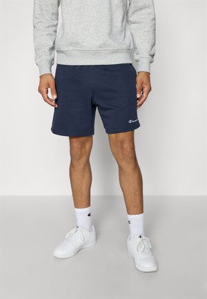 Shorts en coton navy, taille élastique, logo brodé, associés à des baskets blanches et des chaussettes mi-mollet arborant un logo. Sweat-shirt gris visible.