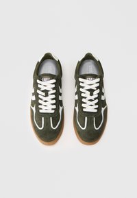 Ein Paar dunkelgrüne Wildledersneaker mit weißen Schnürsenkeln und Akzenten, die Gummisohlen haben, von oben auf einem hellen Hintergrund fotografiert.