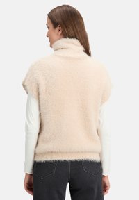 Faux fur gilet in licht beige met een hoge kraag, korte mouwen en een zachte, gestructureerde oppervlakte, gedragen met een witte blouse met lange mouwen.