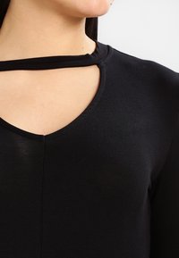 Schwarzes Oberteil mit tailliertem Design, das einen Rundhalsausschnitt und ein halbkreisförmiges Ausschnittdetail am Kragen aufweist. Glatte Textur und dehnbare Stoffqualität.