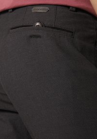 Schwarze Textilhose mit einer Gesäßtasche und Knopfverschluss sowie einem gewebten Logo-Tag. Das Material weist ein dezentes Muster auf, das zusätzliche Details bietet.