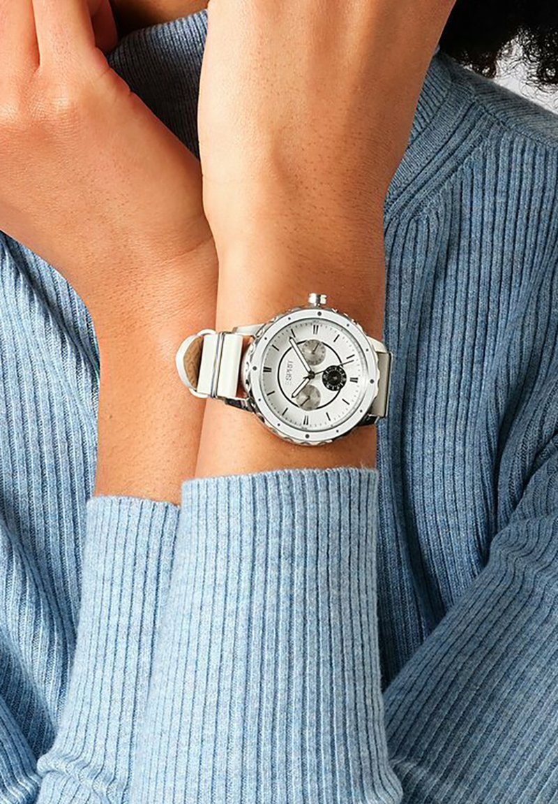 Silberne Armbanduhr mit einem runden weißen Zifferblatt, zwei Unterzifferblättern und einem Metallgliedarmband, getragen an einem Handgelenk über einem blauen gerippten Pullover.
