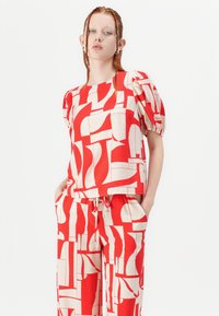 Haut à motif rouge et crème avec des manches bouffantes, associé à un pantalon large assorti. Les tissus semblent légers avec une texture lisse.