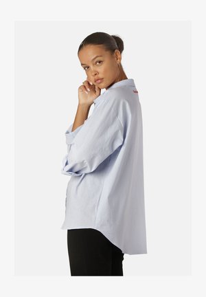 Lichtblauwe oversized blouse met lange mouwen, een gebogen zoom en een rode geborduurde versiering op de achterkant van de kraag. De stof lijkt zacht en gestructureerd.