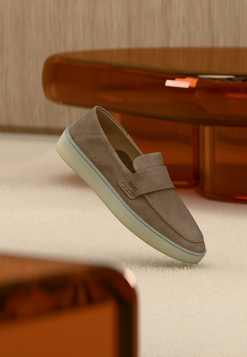 Chaussure en daim marron à enfiler avec une semelle blanche flottant au-dessus d'un tapis beige, avec un mobilier aux tons ambre en arrière-plan.