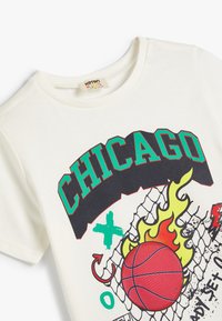 Cremefarvet bomulds t-shirt med et grafisk design, der viser "CHICAGO", en basketball, flammer og et netmønster. Rund halsudskæring, korte ærmer.