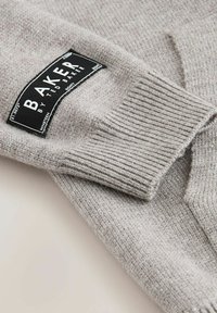 Svetlo šedý pletený sveter s rebrovaným manžetovým zakončením a čiernou nálepkou s bielym textom "BAKER by TED BAKER." Viditeľná mäkká štruktúra.