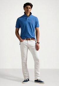 Μπλε πόλο πουκάμισο με γιακά, λευκό τζιν και μπλε αθλητικά παπούτσια. Η εμφάνιση περιλαμβάνει καφέ πλεκτή ζώνη και ρολόι στον καρπό.